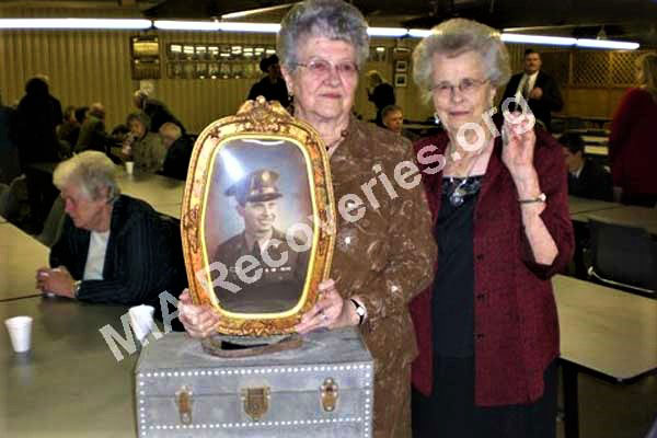 Sisters of Cpl. Arnold B. Stavinoha pose with his portrait and a toolbox he made while home on leave. Viola (in red jacket) holds his recovered ID bracelet in her left hand.